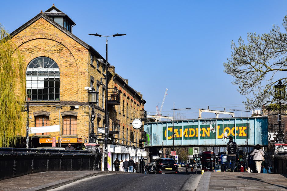 Photograph taken from street level in Bow, London, showing a brick building with large arched window on the upper floor and several smaller windows on the lower floors. A white clock is mounted on a black pole in front of the building, along with streetlights and lamp posts. To the right, a green railway bridge with yellow and green 'Caldon Lock' signage spans across the street, with pedestrians and vehicles including cars and a black van visible below. Nearby, a tree with budding green leaves extends into the frame from the right side, and a few people are walking along the sidewalk, some carrying bags or packages. The scene captures urban street activity related to home relocation logistics, typical of a busy area where moving and furniture transport services, such as those offered by Man With a Van Bow, could operate, with elements of loading processes implied by the setting.