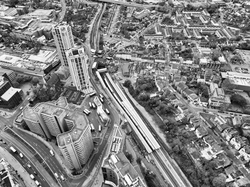Aerial black-and-white photograph of a densely built urban area with high-rise buildings, shorter commercial and residential structures, and a railway line running through the middle. In the foreground, a multi-storey office building is visible, with several vehicles, including buses, parked or moving nearby. The railway tracks run parallel to a road with vehicles, and a train station platform is partially visible along the tracks. The surrounding streets are occupied with parked and moving cars, and numerous residential houses and smaller buildings extend into the background, interspersed with trees and green spaces. The lighting appears natural, highlighting the concentration of infrastructure typical of a city environment. This image may relate to the logistical aspects of home relocation or furniture transport, as exemplified by services like Man With a Van Bow, which handles removals and moving logistics in the area.